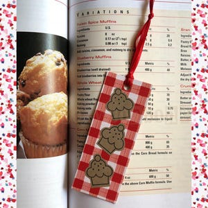 May include: A red and white checkered bookmark with three cupcake designs. The bookmark is placed in an open cookbook with a recipe for muffins. A red ribbon is attached to the top.