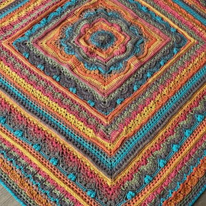 Könnte beinhalten: Eine farbenfrohe, handgefertigte Häkeldecke mit einem zentralen Rautenmuster. Die Decke zeigt eine lebendige Farbpalette, darunter Türkis, Orange, Rosa, Gelb und Grau. Die komplizierten Muster und Texturen schaffen ein gemütliches und dekoratives Textil.
