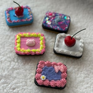May include: Five miniature square cakes with colorful frosting and decorations. One cake has a cherry on top, another has a pig, and another has a bow. The cakes are on a white, textured surface.