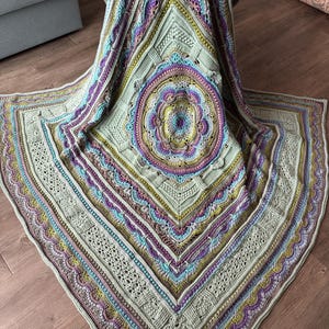 Puede incluir: Una gran manta de ganchillo intrincadamente elaborada en tonos de verde, morado, amarillo y azul. La manta presenta un diseño circular central rodeado de patrones geométricos y florales. Los bordes están acabados con un borde festoneado. La manta se exhibe sobre un suelo de madera.
