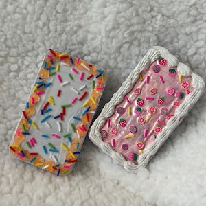 Puede incluir: Dos fundas de teléfono rectangulares y decorativas. Una funda tiene una base blanca con chispas de colores y glaseado naranja y amarillo. La otra funda tiene una base rosa con glaseado blanco y adornos con forma de fruta, como fresas y rodajas de cítricos.