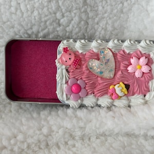 May include: A decorative metal tin with a pink interior and a white and pink frosted top. The top is decorated with a pink bear, a heart, flowers, and a small dessert. The tin is on a white surface.