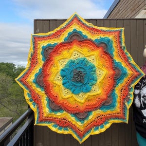 May include: A large, colorful, star-shaped crochet blanket. The blanket features concentric circles of vibrant colors, including yellow, orange, teal, and brown. The intricate design showcases detailed stitching and a textured surface, perfect for home decor.