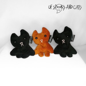 Op de afbeelding: Drie pluche katten speelgoedjes, een zwart, een oranje en een zwart. Elk speelgoedje heeft een klein roze lint op de borst en zwarte ogen. De speelgoedjes zijn in een rij op een wit oppervlak gerangschikt. De tekst "OF SPOOKS AND CATS" is zichtbaar op de achtergrond.