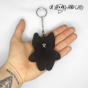 Op de afbeelding: Zwarte pluche katten sleutelhanger met een zilveren ketting en een witte ring. De sleutelhanger wordt vastgehouden in een hand met een tatoeage. De tekst "OF SPOOKS AND CATS" staat boven de kat.