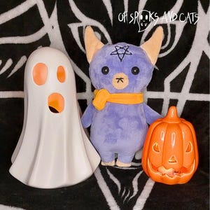 Op de afbeelding: Een Halloween-display met een wit spookbeeldje, een paars pluche speelgoed met een oranje sjaal en een oranje pompoendecoratie. Het pluche speelgoed heeft een pentagram op zijn hoofd. De tekst "OF SPOOKS AND CATS" is zichtbaar.