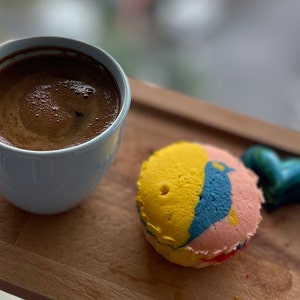 May include: A blue ceramic cup filled with dark coffee sits on a wooden tray with a yellow, blue and pink swirl cake and a green heart-shaped sweet.