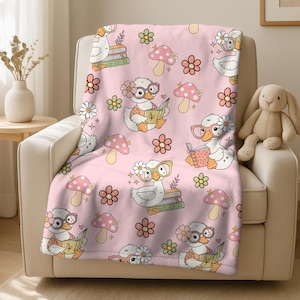 May include: Pink throw blanket featuring a repeating pattern of cartoon ducks wearing glasses and reading books. The design also includes flowers and mushrooms. The blanket is draped over a beige armchair, with a stuffed bunny visible on the right.
