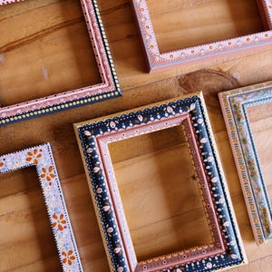 May include: A selection of decorative picture frames, each with a unique hand-painted design. The frames showcase floral and geometric patterns in pink, blue, and navy, with gold detailing. They are displayed on a wooden surface.
