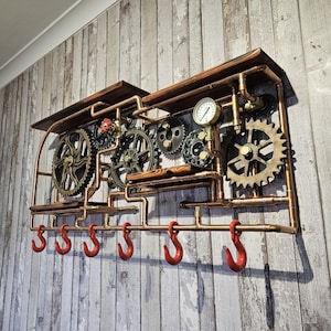 May include: A steampunk-style wall shelf with copper pipes, wooden shelves, and decorative gears. Features a pressure gauge and six red hooks for hanging items. The shelf is mounted on a weathered wood-look wall.