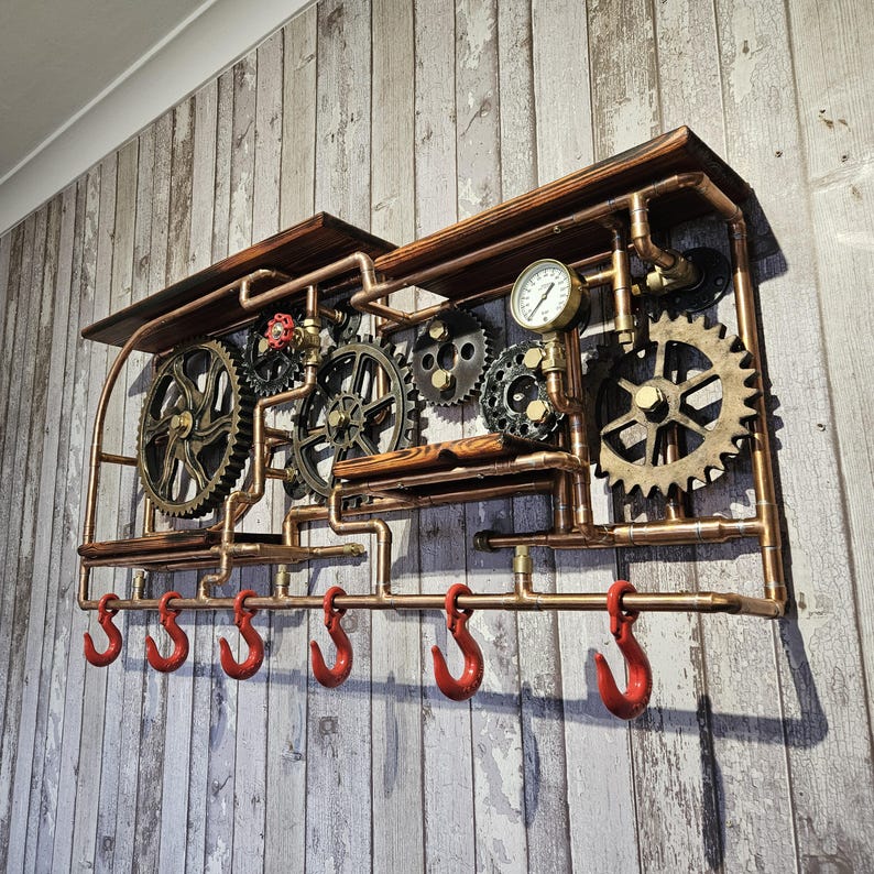 May include: A steampunk-style wall shelf with copper pipes, wooden shelves, and decorative gears. Features a pressure gauge and six red hooks for hanging items. The shelf is mounted on a weathered wood-look wall.