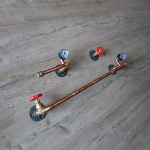 Könnte beinhalten: Wanddekoration und Handtuchhalter aus Kupferrohren. Das Bild zeigt drei Elemente im Industriestil. Zwei haben Manometer, und einer hat einen roten Ventilkurbel. Die Rohre sind auf schwarzen Flanschen montiert.