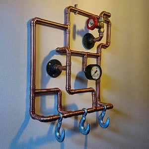 May include: Copper pipe wall mounted coat rack with four hooks and a gauge. The rack is in the shape of a rectangle with a small gauge on the right side.