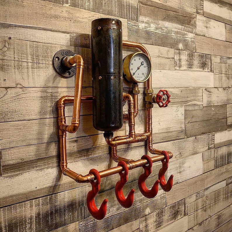 May include: A steampunk-style wall-mounted coat rack made of copper pipes and hooks. The rack features a black metal cylinder, a round gauge, and three red hooks.