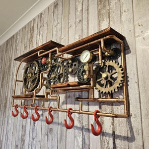 May include: Steampunk-style wall-mounted shelf with copper pipes, gears, and red hooks. The shelf features a pressure gauge and wooden accents, creating an industrial and vintage aesthetic. The shelf is designed for hanging items.
