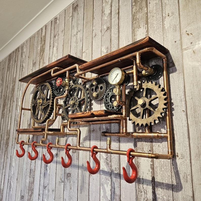 May include: Steampunk-style wall-mounted shelf with copper pipes, gears, and red hooks. The shelf features a pressure gauge and wooden accents, creating an industrial and vintage aesthetic. The shelf is designed for hanging items.