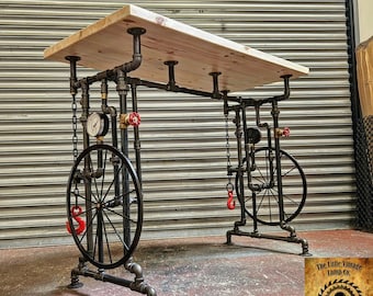 Puede incluir: Una mesa de estilo industrial rústico con una tapa de madera y patas de tubería de metal negro. Las patas presentan un diseño de rueda con cadenas y ganchos rojos. La mesa está etiquetada "The Little Vintage Lamp Co."
