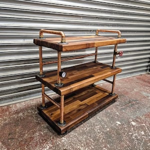 May include: A three-tiered industrial-style cart with a butcher block design. The cart features copper piping and fittings, with wooden shelves. A pressure gauge and a red valve are visible. The cart is designed for storage or display.