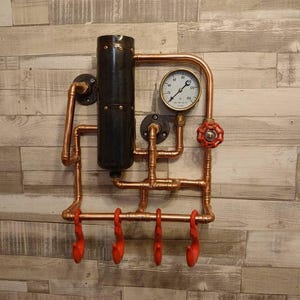 May include: A copper pipe wall mounted coat rack with four red hooks. The rack features a black pipe, a gauge with the numbers 20, 40, 60, 80, 100, 120, and a red valve.