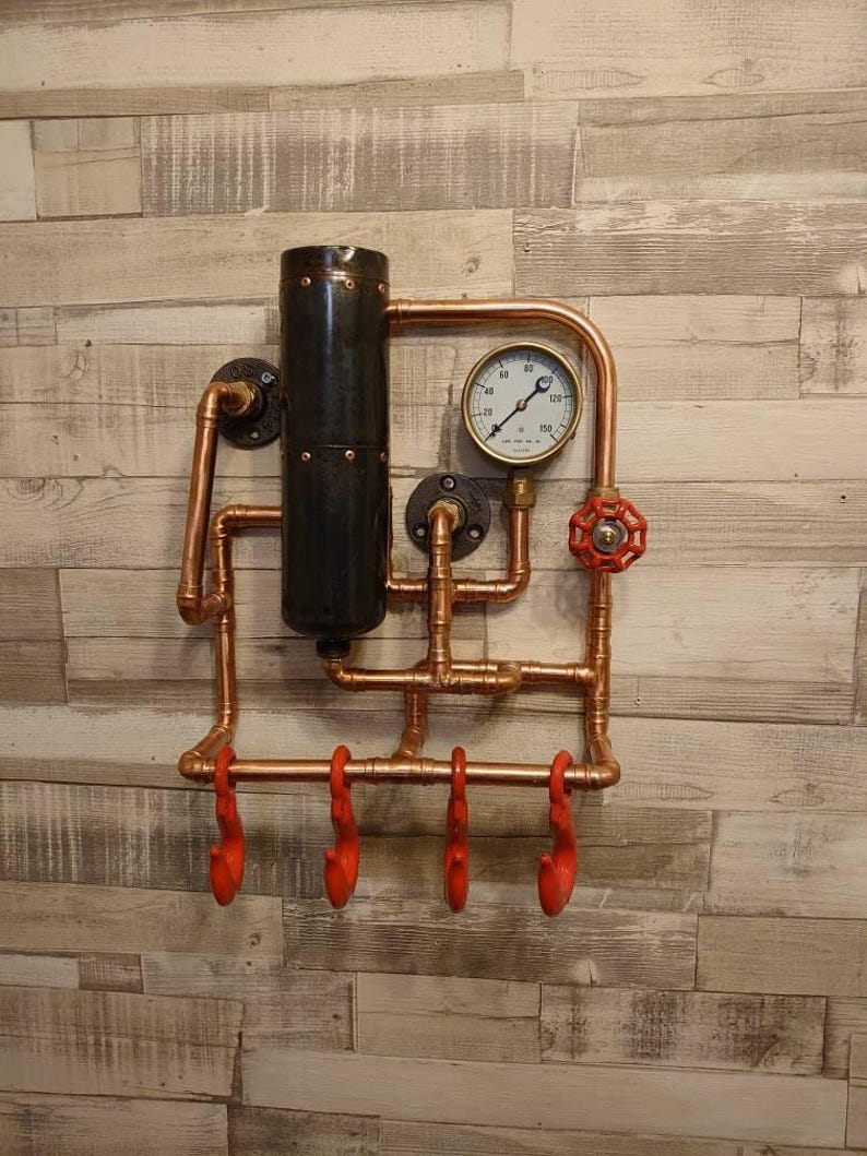 May include: A copper pipe wall mounted coat rack with four red hooks. The rack features a black pipe, a gauge with the numbers 20, 40, 60, 80, 100, 120, and a red valve.