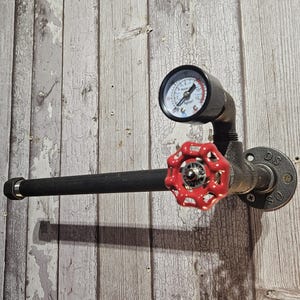 May include: A wall-mounted toilet paper holder constructed from dark gray industrial metal piping. The fixture features a prominent red circular valve handle, a black pressure gauge with a white dial, and a circular mounting flange labeled DS SO.