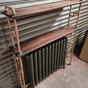 Op de afbeelding: Een tweelaagse wandplank met donkerbruine houten planken, ondersteund door een frame van koperen buizen en fittingen. De constructie is geplaatst over een donkergrijze gietijzeren radiator, waarbij de koperen leidingen doorlopen tot op de vloer.