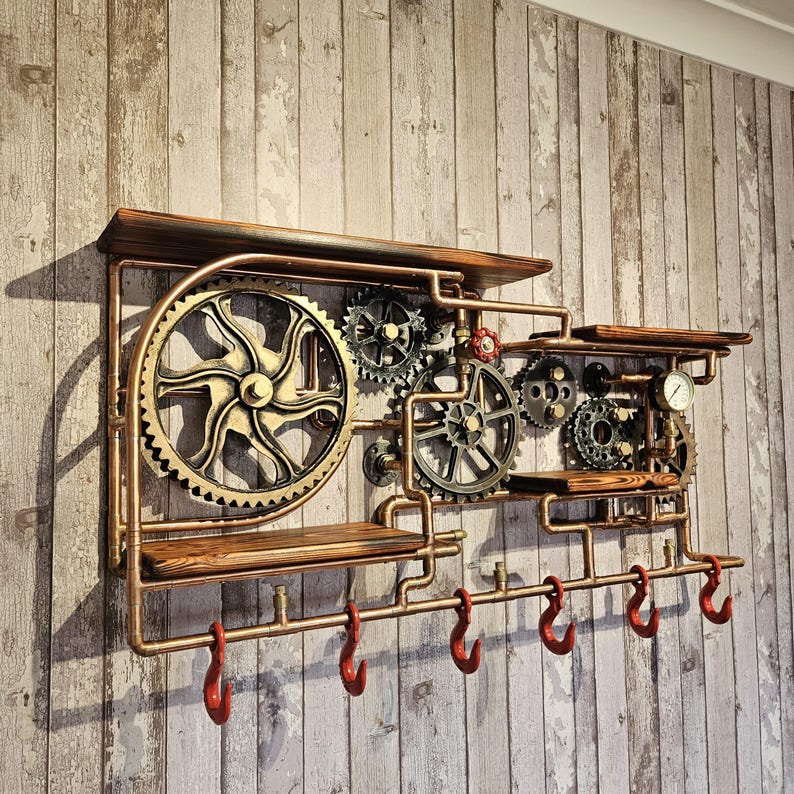 May include: Steampunk-style wall shelf with a copper-colored pipe frame, wooden shelves, and decorative gears. Features six red hooks for hanging items. The design includes a pressure gauge and a red valve.