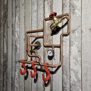 May include: Copper pipe wall art featuring a tap, pressure gauge, and three red hooks. The pipes are arranged in a geometric design, mounted on a distressed wood-effect wall. The tap has a red handle.