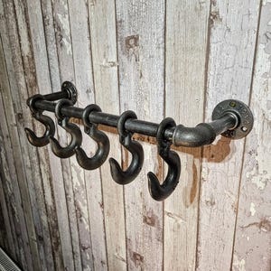 Könnte beinhalten: Ein schwarzer Wand-Kleiderständer im Industriestil mit fünf großen Haken. Der Ständer besteht aus Metallrohren und -fittings mit einem Used-Look. Die Haken sind groß und gebogen, um Mäntel oder andere Gegenstände aufzuhängen. Er ist an einer Holzwand montiert.