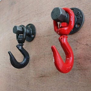 Könnte beinhalten: Zwei Haken im Industriestil, einer schwarz und einer rot, an einer Holzwand montiert. Die Haken sind aus Metall und haben eine abgerundete Form. Sie sind dafür ausgelegt, Gegenstände wie Mäntel, Hüte oder Handtücher zu halten.