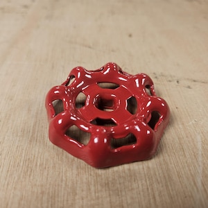 May include: A glossy red metal valve handle featuring a circular design with eight rounded spokes and a square-shaped centre opening. The component has a smooth, reflective finish and a distinct, scalloped outer edge for manual operation.