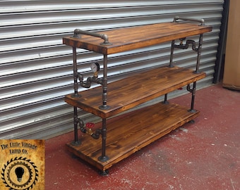 May include: Three-tiered industrial-style shelf unit made of dark brown wood planks and black metal pipes. The top shelf has a metal pipe handle.