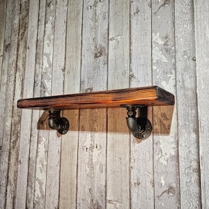 May include: A rustic wooden shelf with a dark stained finish, supported by two black pipe fittings. The shelf is mounted on a weathered, light-colored wooden wall. The shelf is approximately 30 inches long.