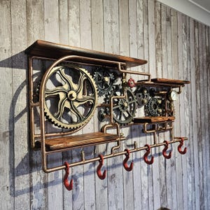 May include: Steampunk-style wall shelf with copper piping, wooden shelves, and decorative gears. Features six red hooks for hanging items. The design incorporates a vintage industrial aesthetic.