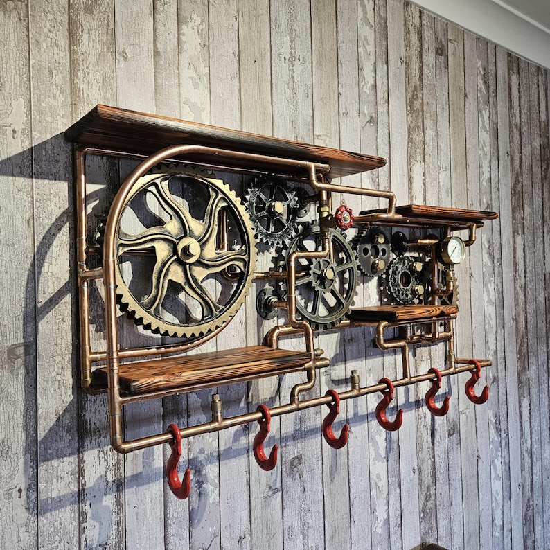 May include: Steampunk-style wall shelf with copper piping, wooden shelves, and decorative gears. Features six red hooks for hanging items. The design incorporates a vintage industrial aesthetic.