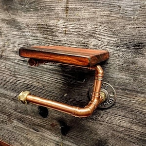 May include: A copper pipe toilet roll holder with a wooden shelf. The holder is mounted to a dark wood wall.