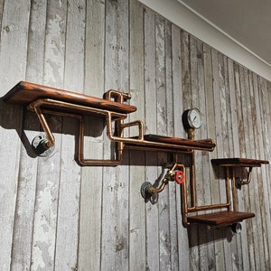 May include: A set of three wooden shelves mounted on a wall, constructed from copper pipes and fittings. The shelves are decorated with a vintage industrial style.