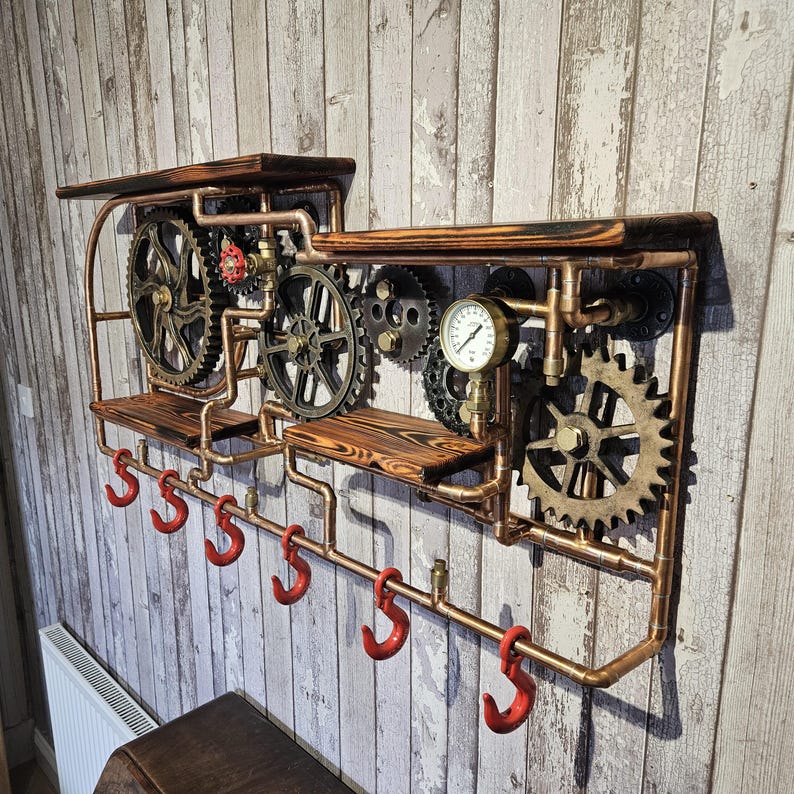 May include: Steampunk-style wall shelf with copper pipes, wooden shelves, and decorative gears. Features red hooks for hanging items. The design includes a pressure gauge and various mechanical components, creating an industrial aesthetic.