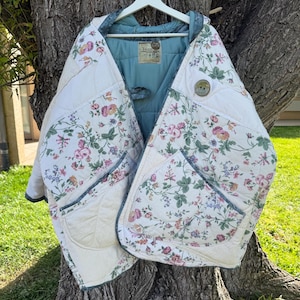 May include: A quilted jacket featuring a cream-colored base with a floral pattern of pink roses and green leaves. The garment has a blue interior lining, a single large button closure, and two front pockets with blue trim edges.