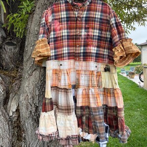 May include: A patchwork dress constructed from multiple plaid flannel shirts in shades of red, orange, brown, and white. The garment features a button-down bodice, ruffled elbow-length sleeves, and a multi-tiered, gathered skirt with raw edges.