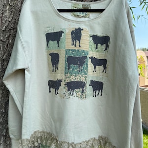 May include: A cream-colored long-sleeve sweatshirt featuring a grid of nine black cow silhouettes printed over various patterned squares. The garment has a scoop neckline and a wide, decorative beige lace trim along the bottom hemline.