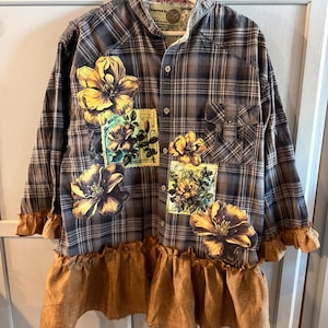 May include: A plaid button-down shirt with large yellow floral appliques and a ruffled hem. The shirt features a mix of brown, gray, and white plaid, with a brown ruffle detail at the sleeves and hem. The shirt is hanging on a white hanger.