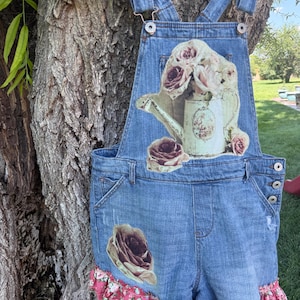 May include: Blue denim short overalls featuring a large graphic print of a watering can filled with roses on the chest. The garment includes a single rose graphic on the left leg and a pink floral patterned ruffle trim along the bottom hem.