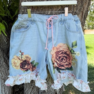 May include: Light blue denim shorts featuring a pink drawstring waist, large floral prints on each leg, and white ruffled hems with a delicate pink and green floral pattern. The shorts are displayed on a white hanger against a tree trunk.