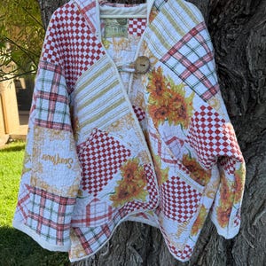 May include: A hooded jacket made from quilted fabric featuring a patchwork design of red and white checkered patterns, green and white plaid, and yellow sunflower prints. The garment includes a single large wooden toggle button closure at the neck.