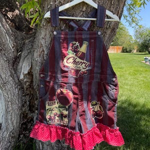 May include: Denim overall shorts with a red and blue striped pattern. The shorts feature cherry-themed graphics, including a "Cherry Cola" logo and "Fresh Berries" text. A red polka-dot ruffle trims the bottom.