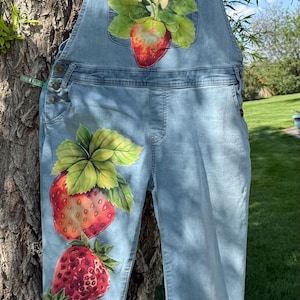 May include: Light blue denim overalls with hand-painted strawberries and flowers. The overalls feature a large strawberry and floral design on the bib and additional strawberry clusters on the legs. The overalls are hanging on a tree.