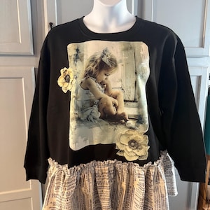 May include: A black long-sleeve sweatshirt featuring a central rectangular graphic of a child sitting near a window. The garment includes two fabric flower appliqués and a ruffled hemline made of beige fabric printed with black alphabet letters.