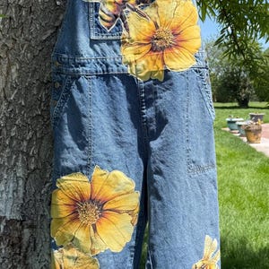 May include: Blue denim overalls featuring large, colorful patches of yellow flowers and honeybees. The bib displays a bee and a large flower, while the legs feature additional floral and bee motifs, including a bee hovering over a honey pot.