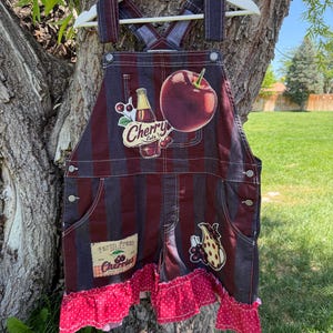 May include: Denim overalls with a red and black striped pattern, featuring cherry-themed patches and a red polka-dot ruffle trim. The overalls have adjustable straps and a front pocket. The patches include a cherry cola bottle, a large red apple, and a cluster of cherries.